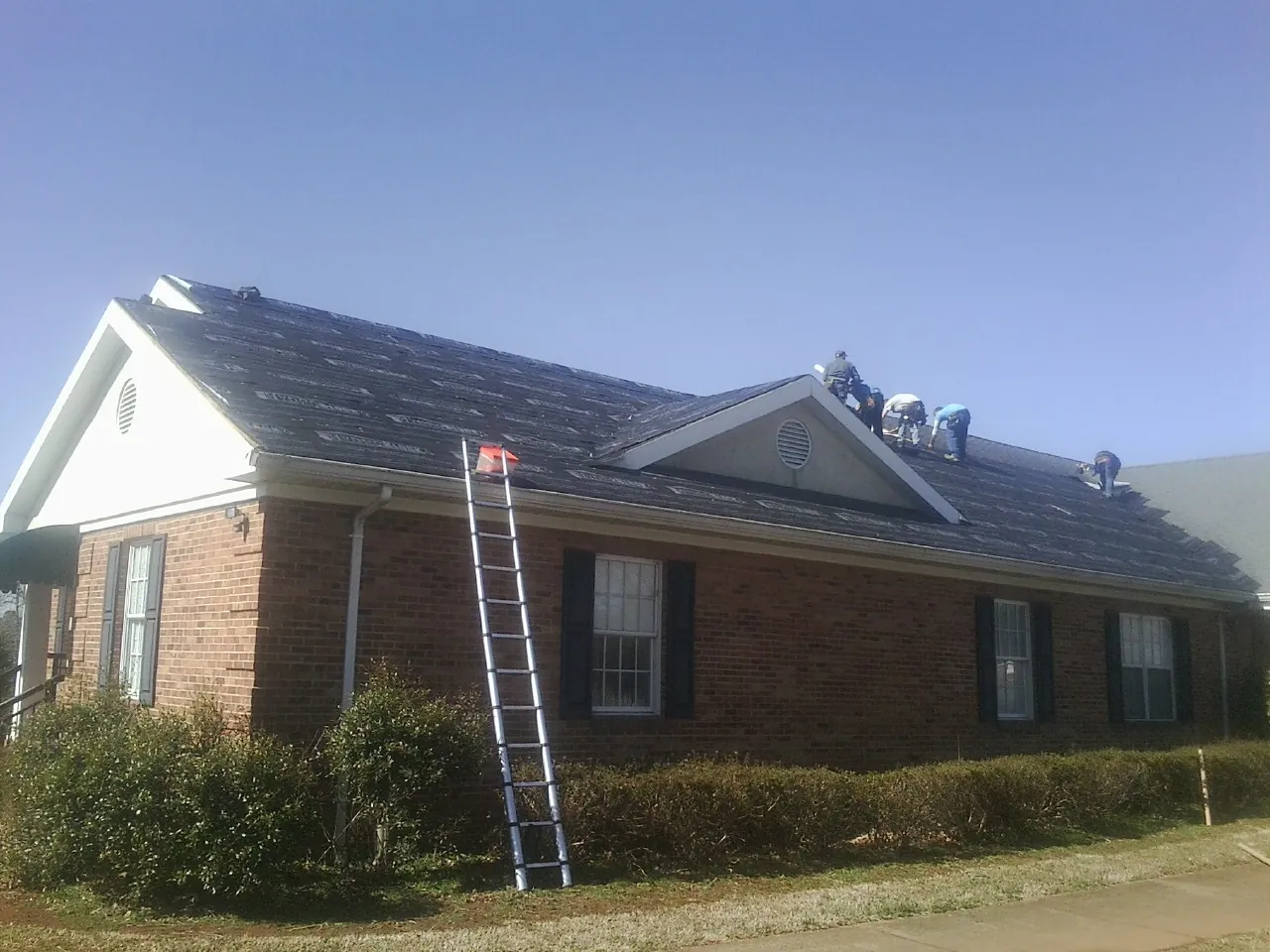 Crew tearing shingles off a brick ranch roof with a ladder set — Emergency Roof Repair work by our crew in Blooming Grove