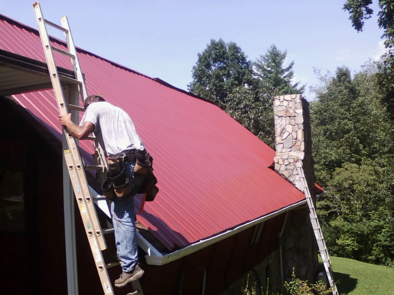 Roofer on a ladder beside a red metal roof and stone chimney — Soffit & Fascia Repair by Roofing Brothers in Blooming Grove